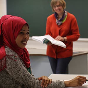 Opiskelija hymyilee ja kouluttaja taustalla hymyilee kirja kädessään.